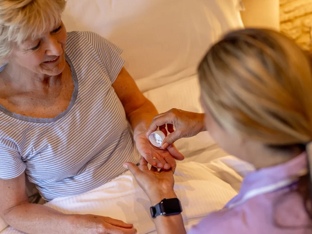 An older woman sitting up in bed smiles as another person hands her medication tablets and a pill bottle, suggesting care or assistance with her health.