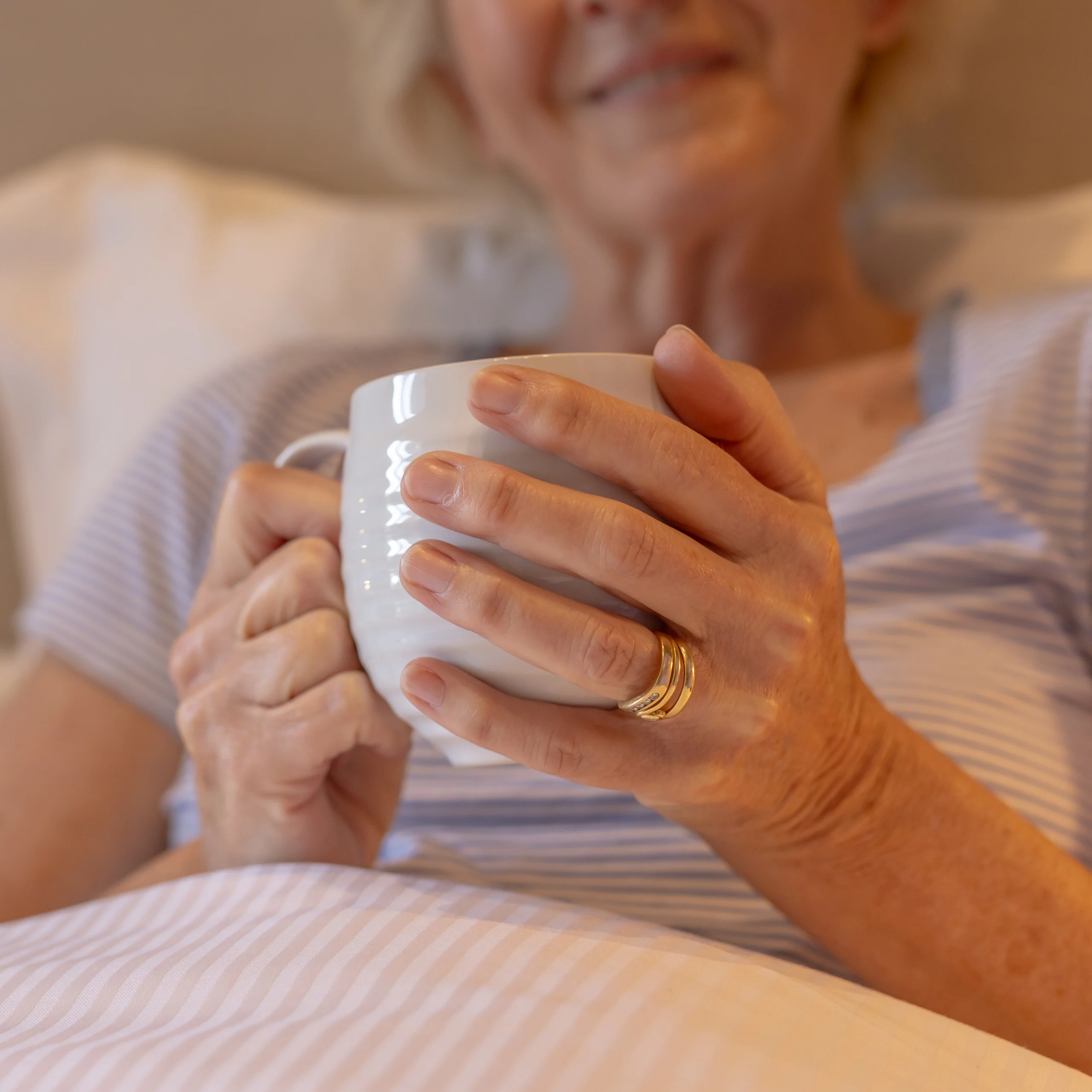 An older woman, partially visible and smiling, relaxes in bed while holding a white mug with both hands. She wears a striped shirt and a gold ring, and the scene appears warm and comforting.