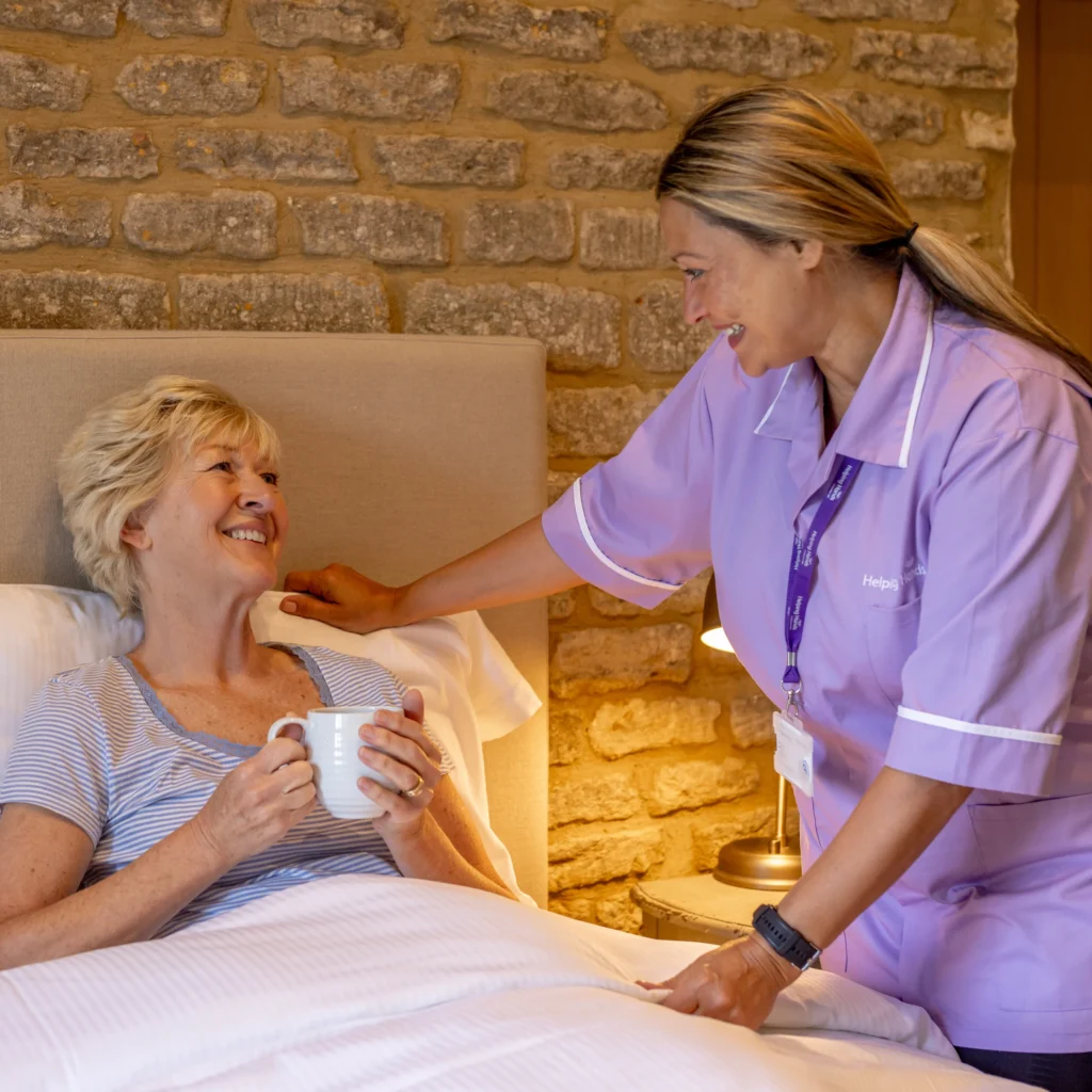A carer in a purple uniform smiles and talks to an elderly woman sitting up in bed, holding a mug, in a cosy room with a stone wall background.
