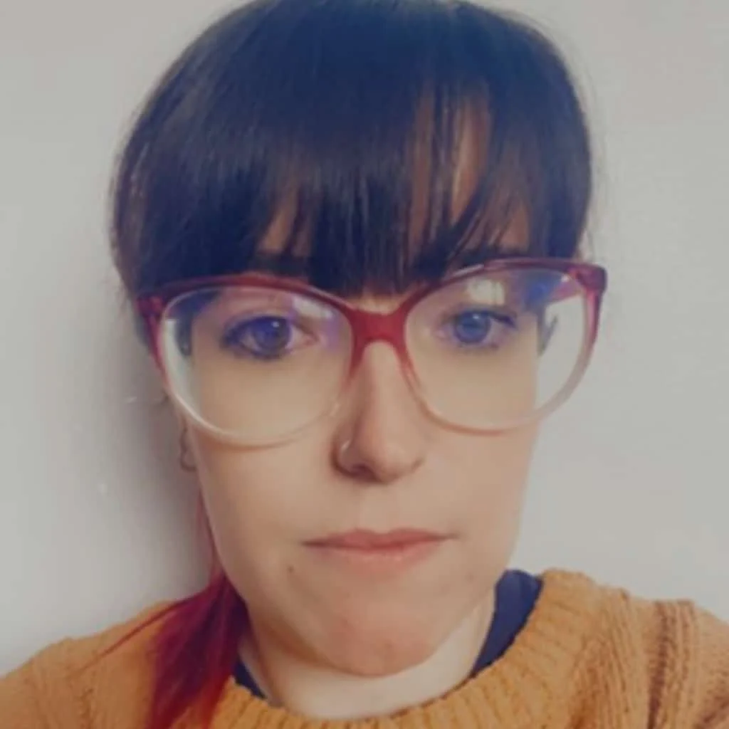 A person with long dark hair and fringe, wearing large, round glasses with red frames and a brown jumper, is looking directly at the camera against a plain light background.