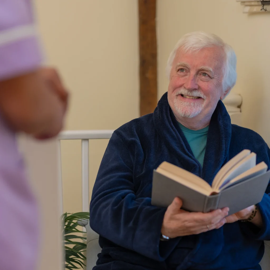 An older man with white hair and a beard, wearing a dark blue dressing gown, sits and smiles while holding an open book. He looks up at a person standing nearby, who is partially visible.