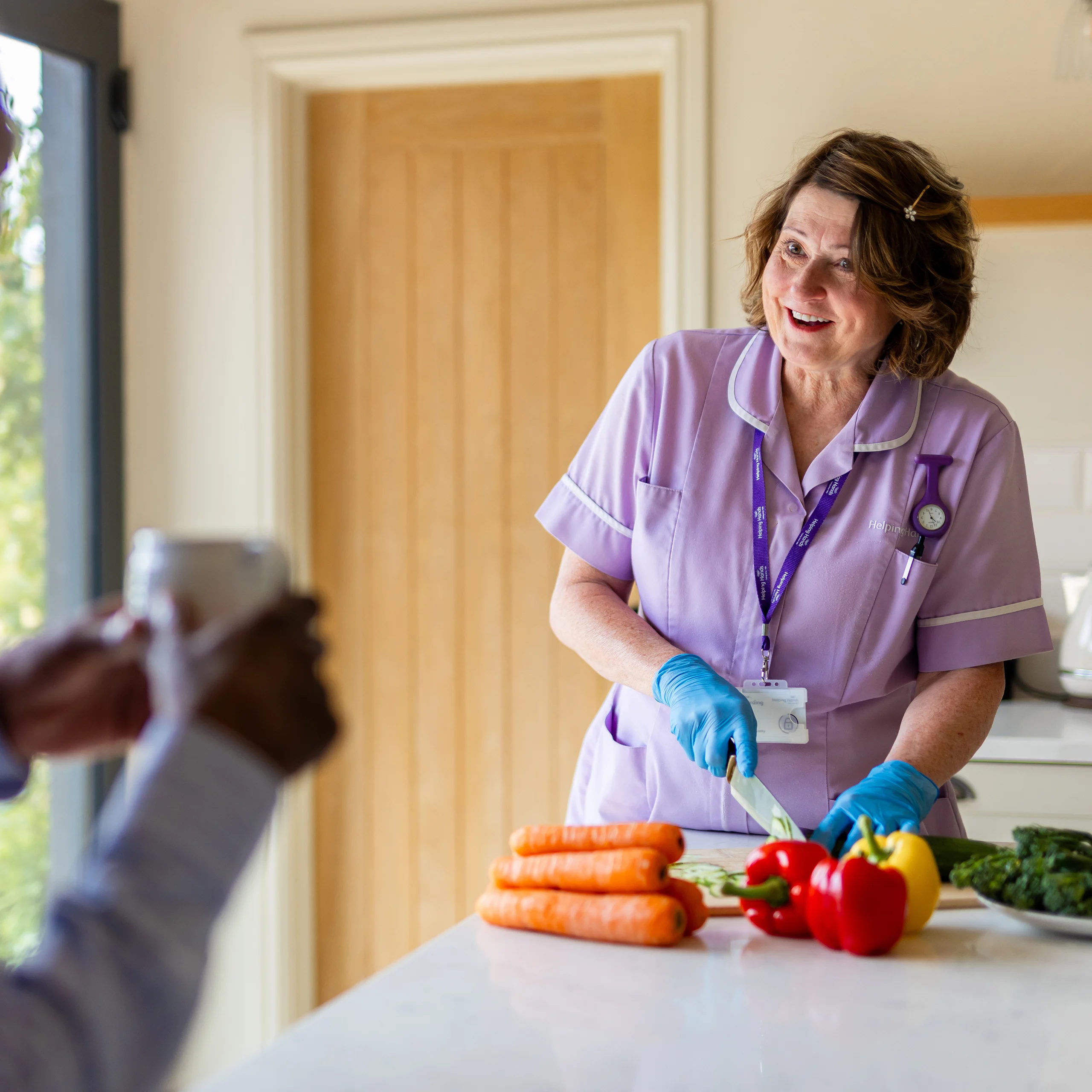 A smiling carer in a purple uniform and blue gloves slices vegetables, including carrots and peppers, on a kitchen worktop while talking to another person holding a mug.