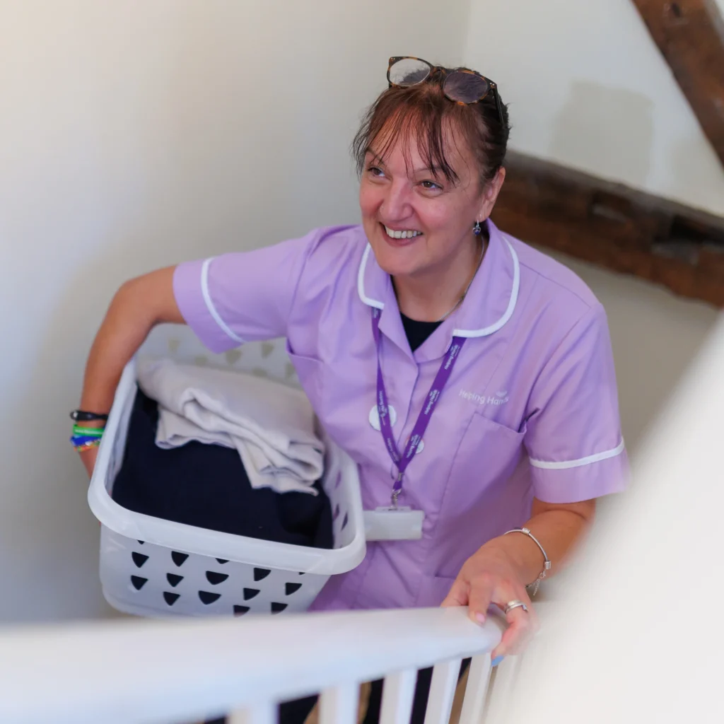 A smiling woman in a light purple uniform carries a laundry basket with folded clothes up a staircase, wearing an ID badge and glasses on her head.