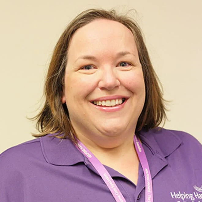 A woman with shoulder-length brown hair smiles at the camera. She is wearing a purple polo shirt and a matching lanyard. The background is a plain, light-coloured wall.