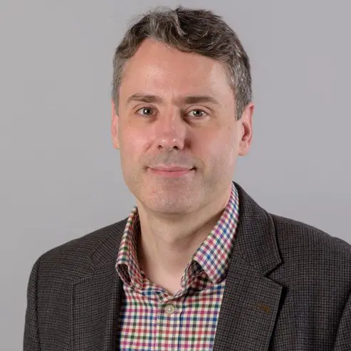 A man with short grey hair wearing a brown blazer and a checked shirt, smiling softly, poses in front of a plain grey background.