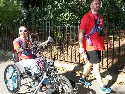 Two people wearing red shirts and medals are outdoors on a pavement. One is using a handcycle and smiling, while the other is walking beside him with a camera around his neck. Trees and a black fence are in the background.
