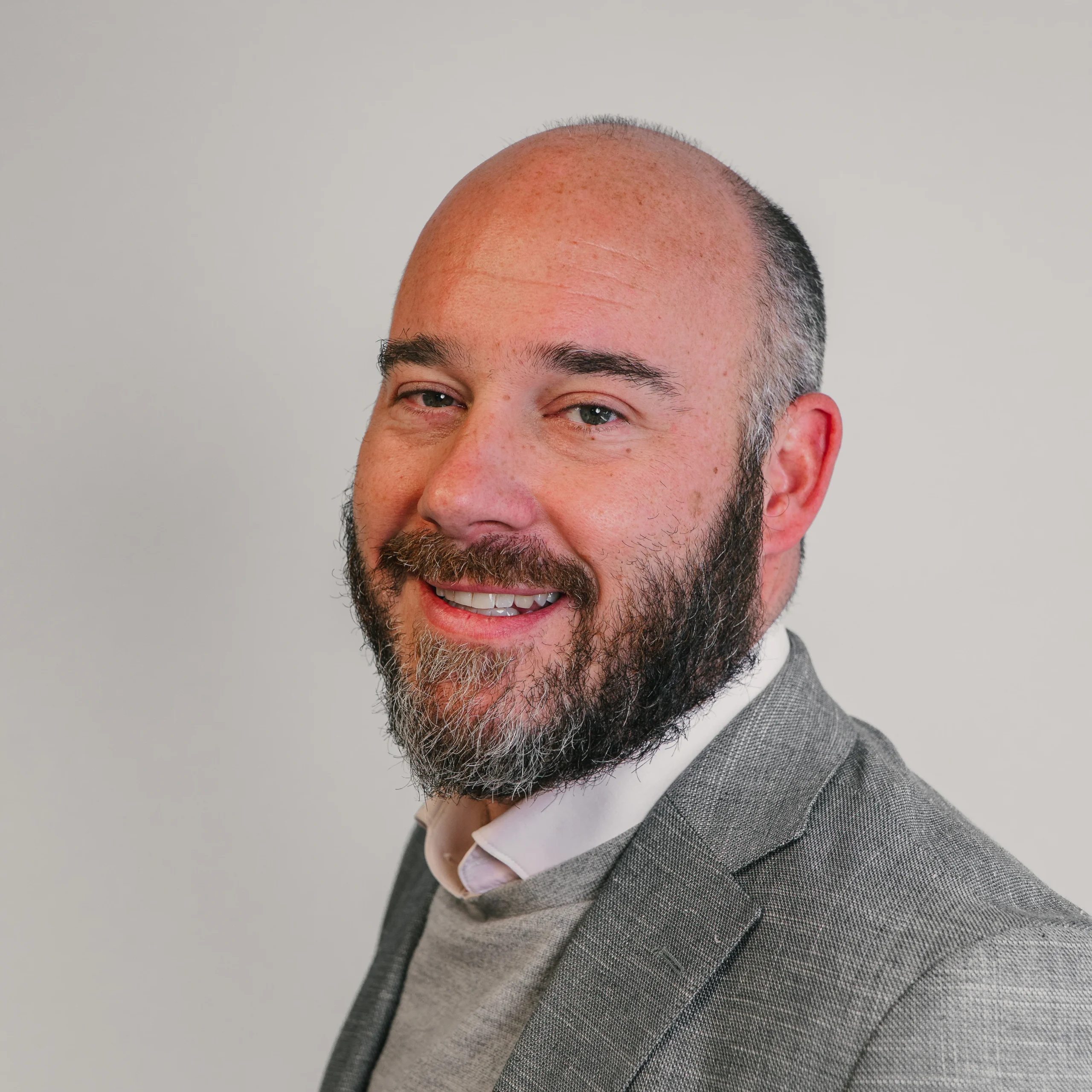 A man with a bald head and full beard, wearing a grey suit jacket over a light jumper and white shirt, smiles at the camera against a plain light background.