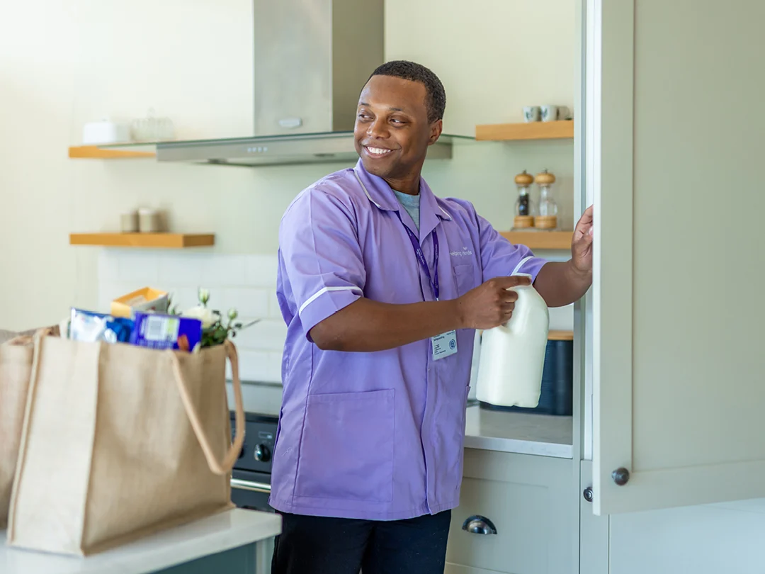 A man in a purple uniform smiles whilst storing a bottle of milk in a kitchen cupboard; a shopping bag of food sits on the counter beside him.
