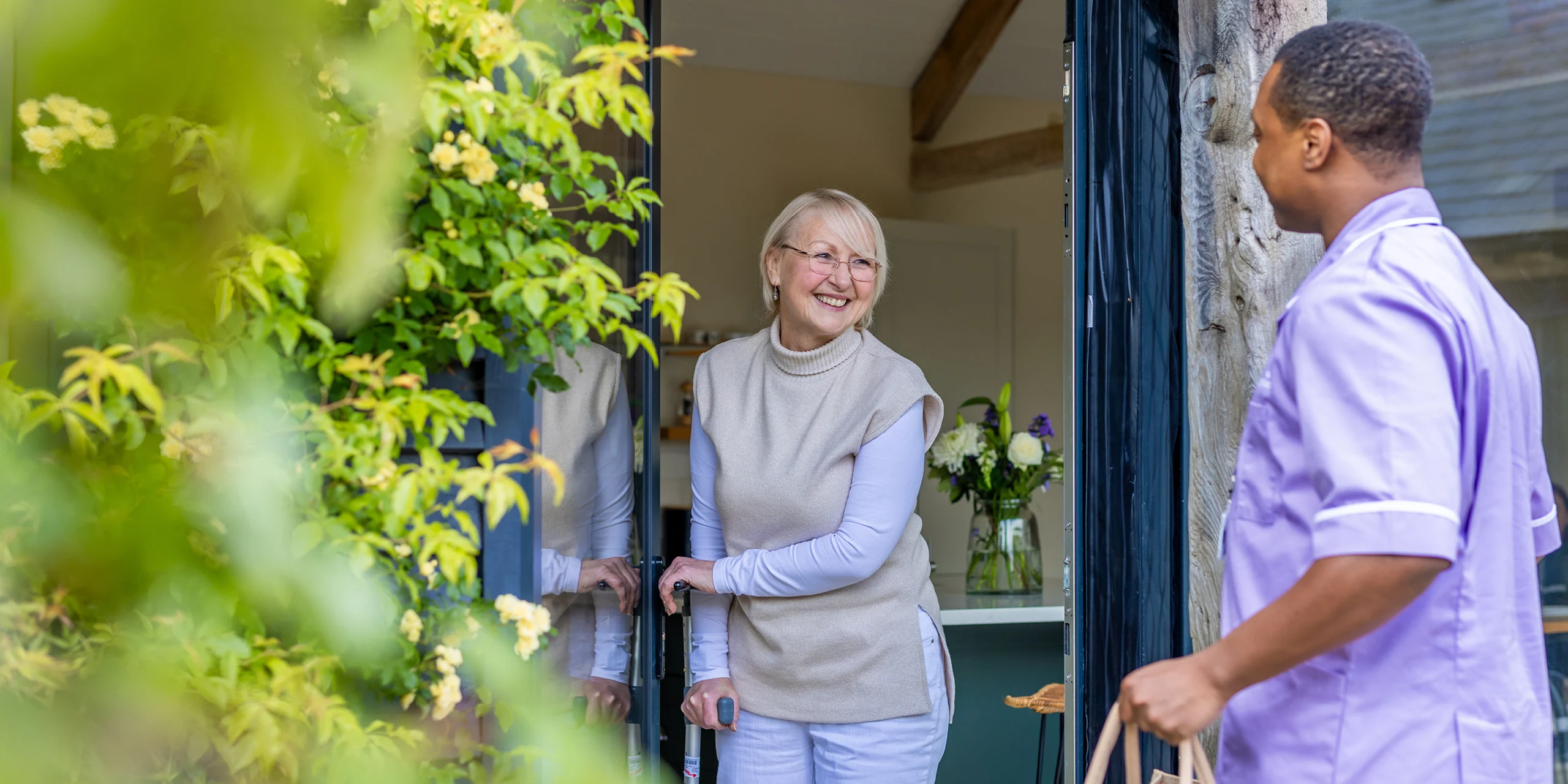 An older woman with a walking stick stands smiling at her open door, while a person in a light purple uniform offers her a paper bag. Green leaves frame the foreground.