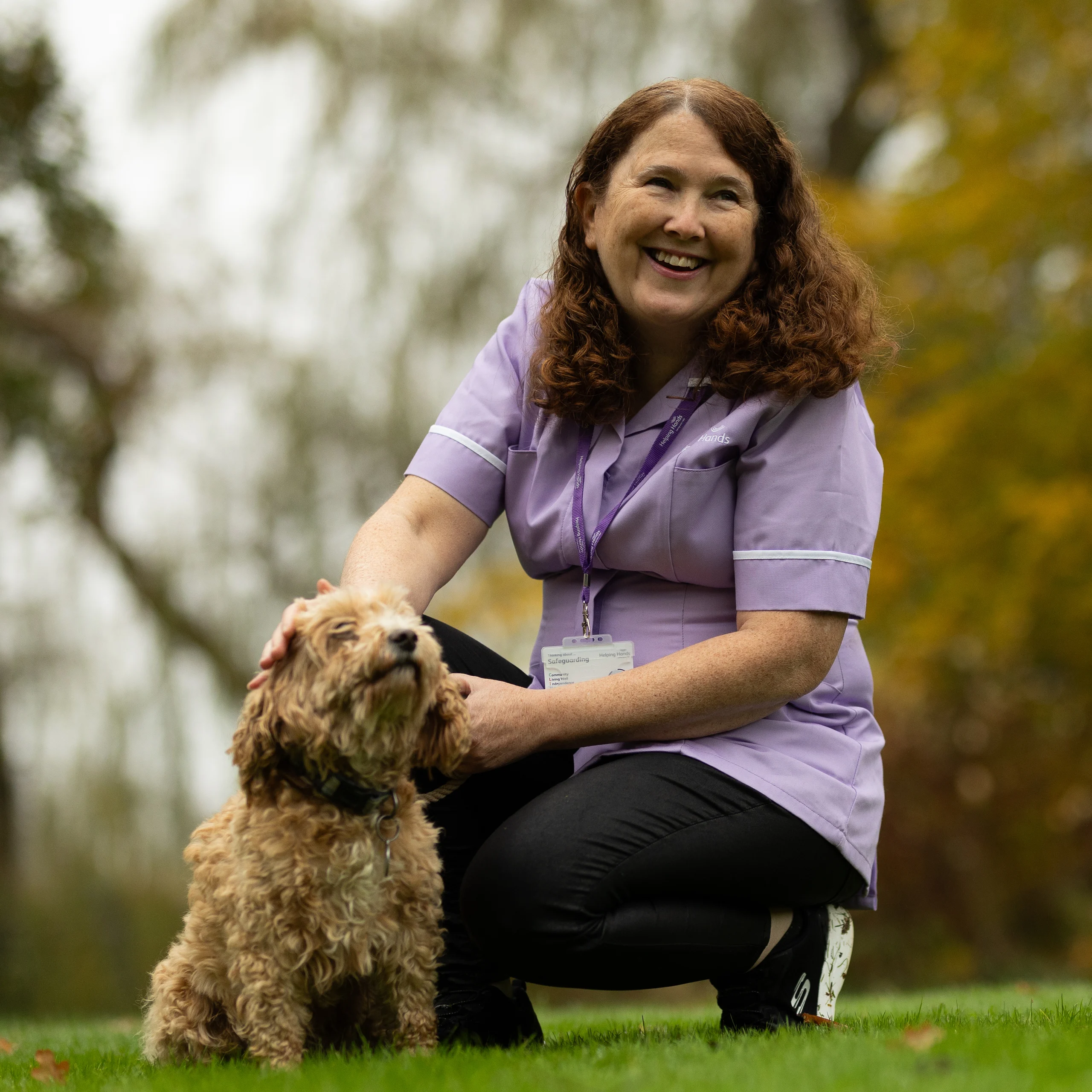 A woman in a purple uniform smiles and kneels on the grass, stroking a small, curly-haired dog. Trees with autumn leaves are blurred in the background.