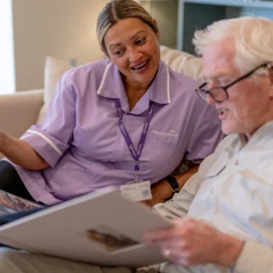 A smiling carer in a purple uniform sits beside an elderly man with white hair and glasses, looking at a large photo album together in a comfortable, well-lit room.