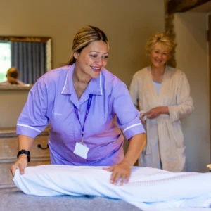 A carer in a purple uniform smiles whilst making a bed, as an older woman in a dressing gown stands nearby watching and smiling in a cosy, warmly lit room.