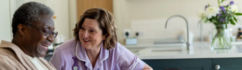 A nurse in a light purple uniform smiles whilst talking to an elderly man in a kitchen with flowers on the counter in the background.