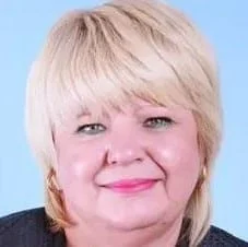 A middle-aged woman with short blonde hair, light green eyes, and pink lipstick, smiling gently at the camera against a light blue background.