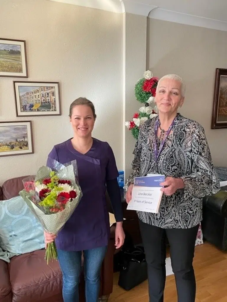 Two women stand indoors, one holding a bouquet of flowers and the other holding a certificate. Both are smiling. Behind them are framed pictures on the wall and a decorative flower arrangement.