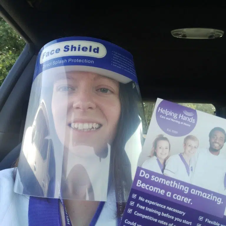 A smiling person wearing a face visor sits in a car and holds a Helping Hands leaflet promoting care jobs, featuring photos of diverse carers and a list of job benefits.