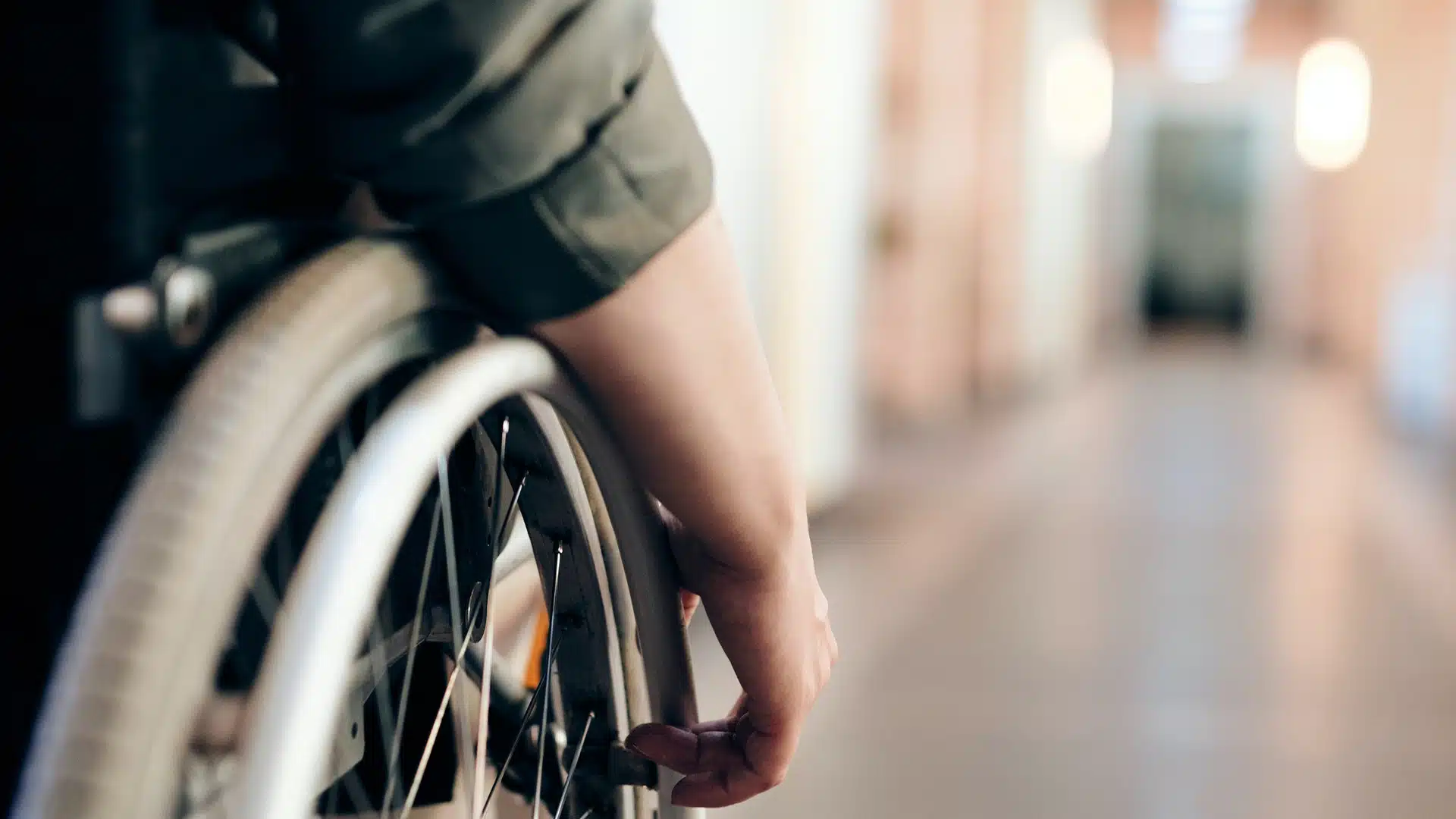 A close-up of a person’s hand on the wheel of a wheelchair in a corridor, with the background softly blurred.