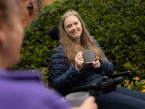 A woman in a wheelchair, wearing a blue jacket, smiles whilst holding a mug outdoors. Another person, slightly out of focus, also holds a mug in the foreground. Green bushes and a brick wall are in the background.