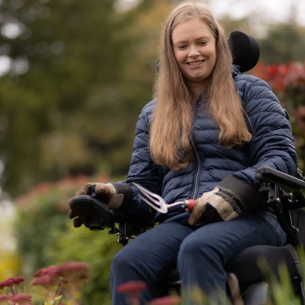 A young woman with long blonde hair and a blue jacket sits in a wheelchair outdoors, smiling and holding a gardening tool. She is surrounded by flowers and greenery.