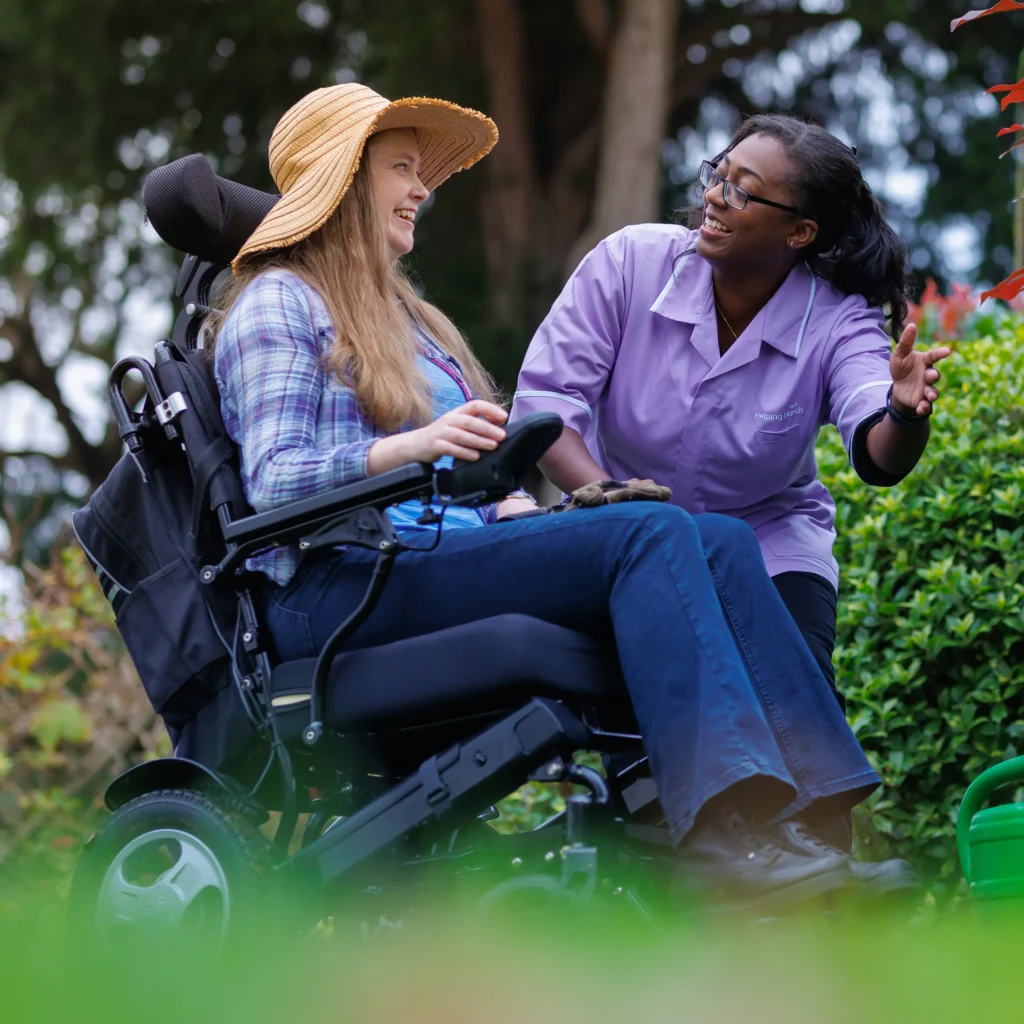 A woman in a wheelchair wearing a sunhat smiles and talks with a carer in a purple shirt. They are outdoors in a garden, surrounded by greenery and flowers.