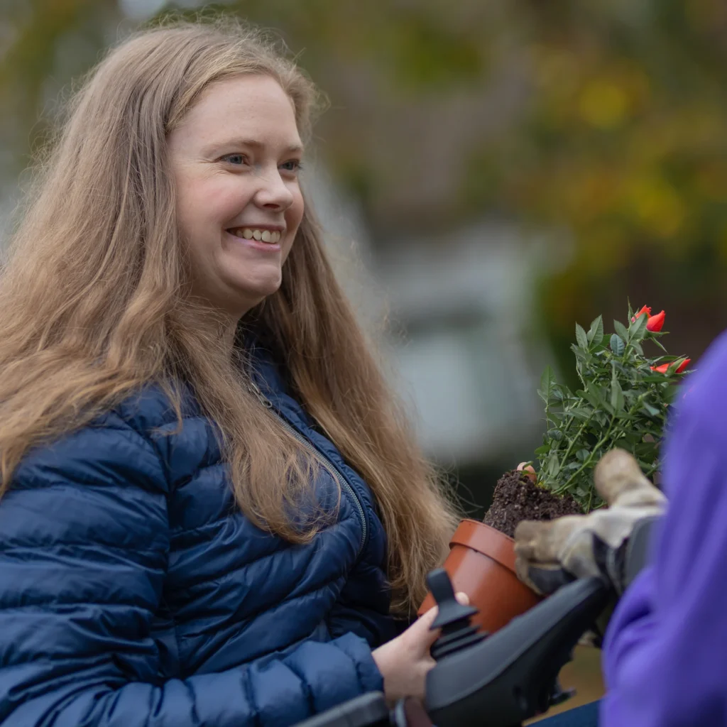 A smiling woman in a blue jacket holds a small potted plant with red flowers, handing it to another person wearing gardening gloves. The background is outdoors and blurred.